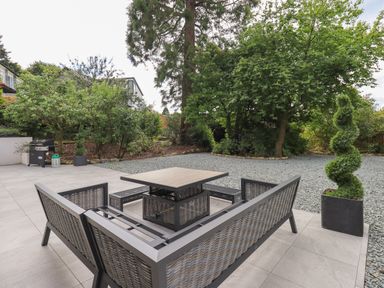 An outdoor patio with grey tiled flooring a woven seating set with a square table a spiral shaped topiary and trees at Hares Breath in Bowness-On-Windermere