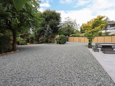 An outdoor area with a gravel ground surrounded by trees and a fence with a patio area containing a table and benches at Hares Breath in Bowness-On-Windermere