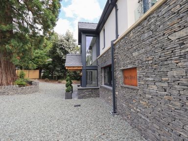 The exterior of a house with stone walls a gravel driveway and trees at Hares Breath in Bowness-On-Windermere
