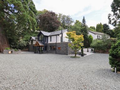 A modern two-story house with stone and white exterior surrounded by trees and a gravel driveway at Hare's Breath in Bowness-On-Windermere