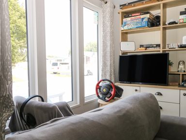 A living room with a sofa a television a gaming steering wheel and board games on shelves at The Goodwood Lodge in Runcton near Chichester
