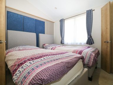 A bedroom with two single beds lined up side by side with patterned bedding a window with blue patterned curtains and wooden doors at The Goodwood Lodge in Runcton near Chichester