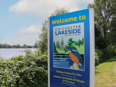 A welcome sign for Chichester Lakeside Holiday Park near a body of water with trees and bushes in the background