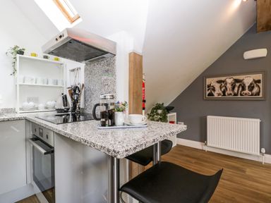 A kitchen with a granite countertop, black bar stools, a stove, kettle, and a framed cow painting on a gray wall at The Coach House in Ludlow