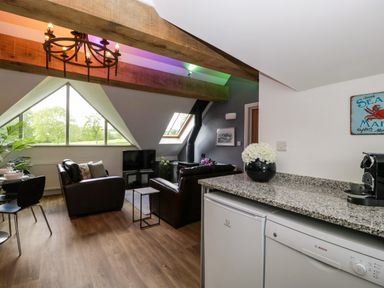 A living room with brown leather sofas dining table under a large triangular window and kitchen counter with appliances at The Coach House in Ludlow