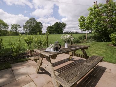 A wooden picnic table with a coffee press and flowers on a patio overlooking a green field at The Coach House in Ludlow