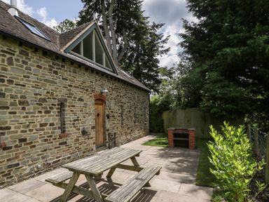 An outdoor patio area with a wooden picnic table a brick barbecue against a stone house with trees around at The Coach House in Ludlow