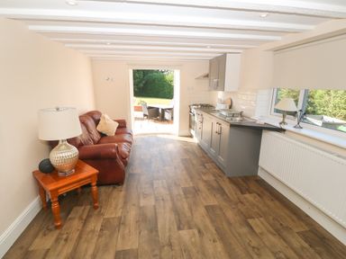 A kitchen and living room with a brown leather sofa wooden side table with a lamp kitchen cabinets and a window at Plas Eithin in Mynydd Mechell near Llanfechell