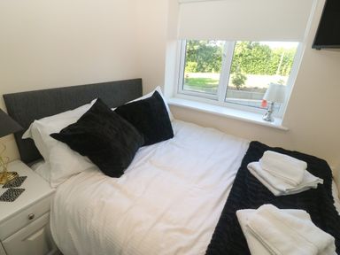 A bedroom with a bed with white sheets and black pillows beside a window with a lamp on the windowsill at Plas Eithin in Mynydd Mechell near Llanfechell