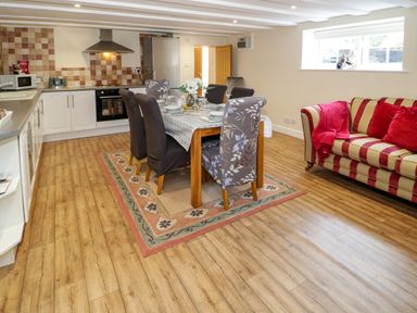 An open plan kitchen and dining area with a wooden table and six chairs on a rug beside a striped sofa with red cushions at Plas Eithin in Mynydd Mechell near Llanfechell