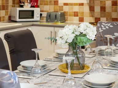 A dining table set with white plates and upside down wine glasses with a vase of white flowers in a kitchen at Plas Eithin in Mynydd Mechell near Llanfechell