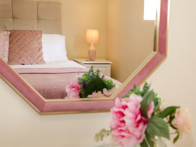 A bedroom with a bed with cushions and a lamp on a bedside table reflected in a mirror with flowers in front of it at Plas Eithin in Mynydd Mechell near Llanfechell