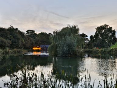 A pond with a small island and trees surrounding it at Oak Lodge At Bridge Lake Farm & Fishery in Chacombe