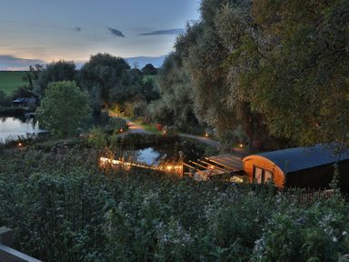 A landscape with a pond a small path trees and a wooden cabin at Oak Lodge At Bridge Lake Farm & Fishery in Chacombe