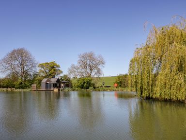 A building by a lake surrounded by trees at Oak Lodge At Bridge Lake Farm & Fishery Chacombe