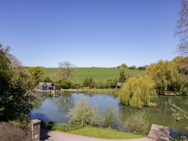 A view of a lake with greenery and a building at Oak Lodge At Bridge Lake Farm & Fishery Chacombe