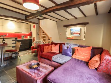 A living room with a multicolored sofa and pillows a small glass dining table wooden stairs and kitchen area with red backsplash at The Curious Orange in Troutbeck