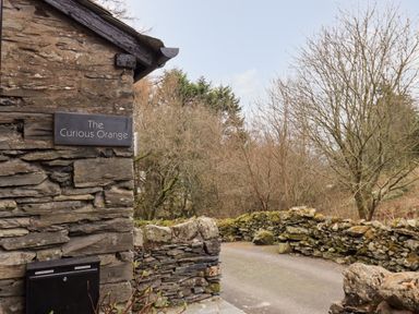 A stone wall with a sign that says The Curious Orange and a narrow road with stone fences and leafless trees at The Curious Orange in Troutbeck