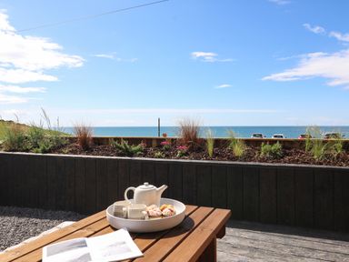 A wooden table with a teapot cups and pastries with a view of the sea and plants at Sandpiper Chalet in Eype