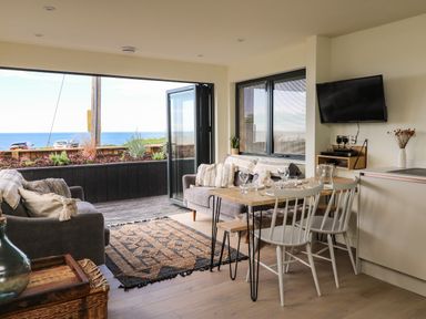 A living room with sofas and a dining table near an open door overlooking the sea at Sandpiper Chalet in Eype