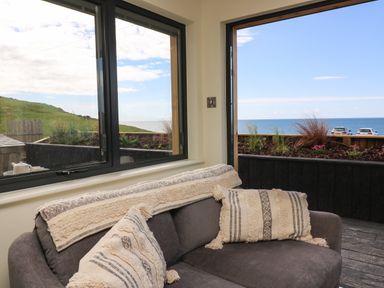 A grey sofa with cream textured cushions near windows overlooking a garden and the sea at Sandpiper Chalet in Eype
