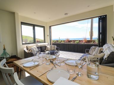 A dining table set with plates and glasses next to a seating area with sofas by large open windows showing an outdoor ocean view at Sandpiper Chalet in Eype