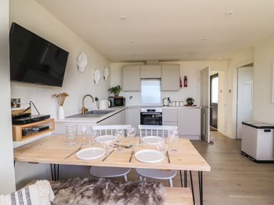 A kitchen and dining area with a wooden table set with plates and glasses and kitchen cabinets in the background at Sandpiper Chalet in Eype