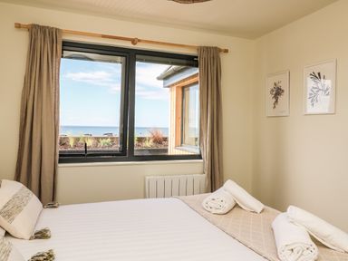 A bedroom with a bed with pillows and rolled towels near a window with beige curtains and sea view at Sandpiper Chalet in Eype