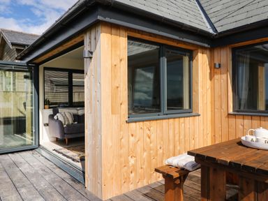 An outdoor wooden deck with a wooden picnic table and bench and an indoor living room with a sofa and cushions at Sandpiper Chalet in Eype