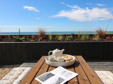An outdoor wooden picnic table with teapot, cups, snacks, and an open book with ocean view in the background at Sandpiper Chalet in Eype