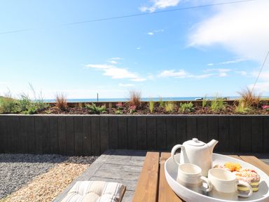An outdoor wooden table with a tray holding a teapot two mugs and cupcakes with a planter and blue sky in the background at Sandpiper Chalet in Eype