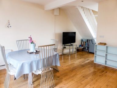 A dining area with a round table covered with a blue polka dot tablecloth and metal chairs beside a living space with a TV on a glass stand and a small bookshelf under a staircase at Beach View in Duncannon near Ramsgrange County Wexford