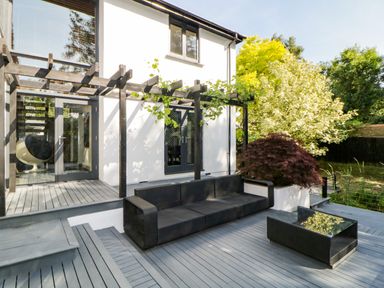 An outdoor patio with black wicker sofa and table next to a white house with wooden pergola and plants at Iroko in Millbrook