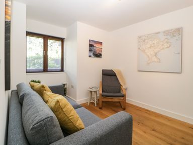 A small sitting room with a gray sofa and yellow cushions a wooden floor a gray armchair with a beige throw a side table books a window and wall art at Iroko in Millbrook