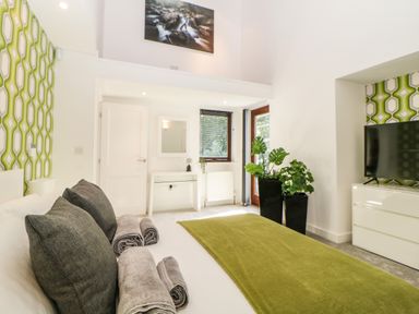 A bedroom with a bed with green throw and grey pillows and towels two black planters with green plants white dresser with a television and patterned green wallpaper at Iroko in Millbrook