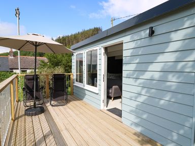 A wooden deck with two black chairs under a beige umbrella outside a light blue building at Caban Glas in Llanafan near Pontrhydfendigaid