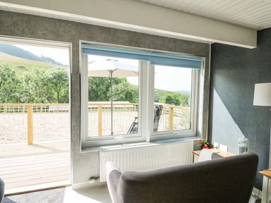 A room with a gray sofa facing windows and a door leading to a wooden deck with an umbrella and chairs at Caban Glas in Llanafan near Pontrhydfendigaid
