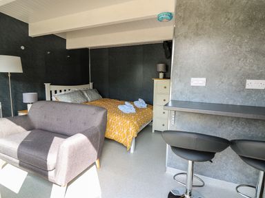 A bedroom with a bed with yellow and gray bedding a gray sofa a floor lamp a chest of drawers and two black bar stools at Caban Glas in Llanafan near Pontrhydfendigaid