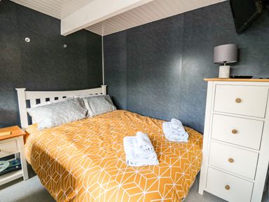 A bedroom with a bed with orange patterned bedspread two folded towels white bedside table and white dresser with lamp at Caban Glas Llanafan near Pontrhydfendigaid