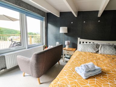 A bedroom with a bed with yellow patterned bedspread and towels on it a gray sofa near a window and bedside tables with lamps at Caban Glas in Llanafan near Pontrhydfendigaid