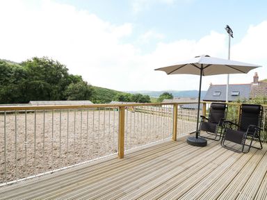 A wooden deck with two black chairs and a large umbrella overlooking a fenced outdoor area and hills at Caban Glas Llanafan near Pontrhydfendigaid