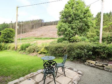 A small outdoor patio with a metal table and two chairs facing a grassy area and trees at Caban Glas in Llanafan near Pontrhydfendigaid