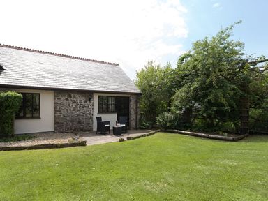 A cottage with a stone and white exterior a small patio with two chairs and a table and a green lawn with trees at Honey Bee Cottage in Bradworthy