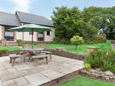 An outdoor patio with wooden picnic table and umbrella near a stone wall and garden at Honey Bee Cottage in Bradworthy