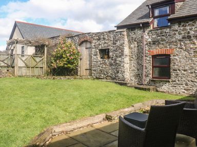 A garden with grass a stone wall and wooden door next to a stone house with wicker chairs on a patio at Honey Bee Cottage in Bradworthy