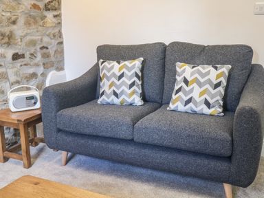 A grey sofa with two patterned cushions next to a wooden side table with a small radio on it at Honey Bee Cottage in Bradworthy