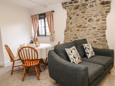 A dining area with a round table and wooden chairs next to a dark gray sofa with patterned cushions by a stone wall at Honey Bee Cottage in Bradworthy