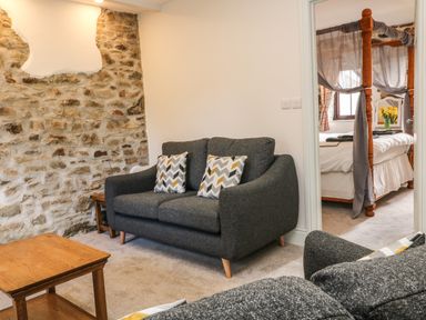A sitting area with grey sofas and patterned cushions next to a stone wall and a wooden coffee table with a view into a bedroom at Honey Bee Cottage in Bradworthy