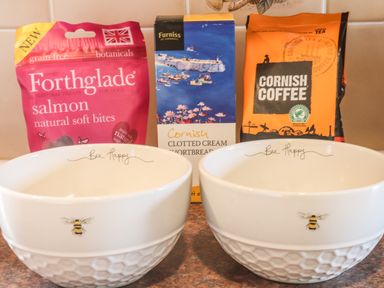 Two white bowls with bee designs and the text Bee Happy in front of dog treats, clotted cream shortbread, and Cornish coffee packets on a kitchen counter