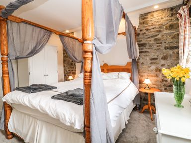 A bedroom with a four-poster bed stone walls nightstand with lamp wardrobe and flowers on a table at Honey Bee Cottage in Bradworthy